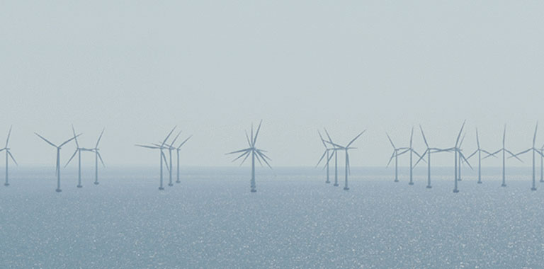 Ausschnitt eines Offshore Windparks. Zu sehen sind ca. 20 Windkrafträder. Das Wetter ist eher diesig und das Meer ist ruhig.